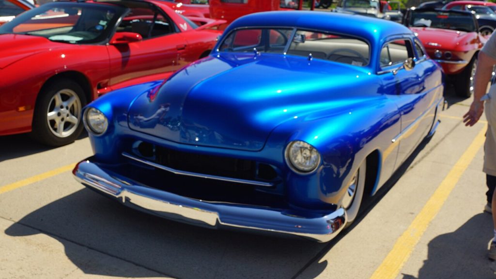Custom metallic blue 1950 Mercury Lead Sled with chopped roof, chrome bumper and lowered stance at car show beside red vehicles