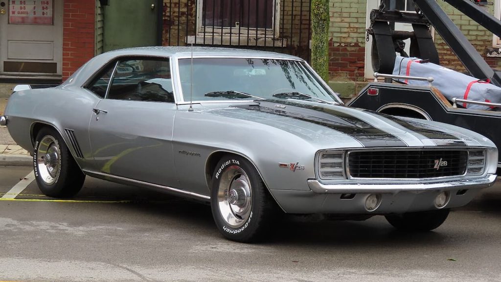 Silver 1969 Chevrolet Camaro Z28 with black racing stripes parked on street in front of brick buildings