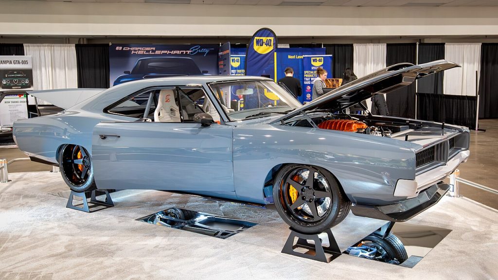 Light blue classic 1969 Dodge Charger Hellephant with open hood with Mopar Custom Stage 3 Crate HEMI displayed on wheel stands at indoor automotive show or convention