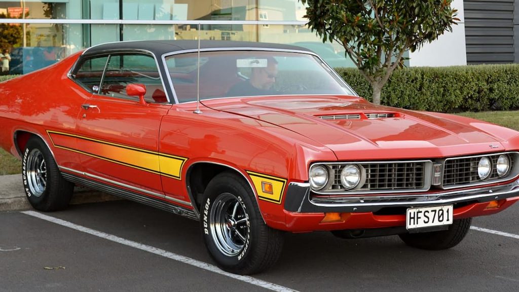 Red 1971 Ford Torino muscle car with yellow racing stripes parked in lot outside modern glass building