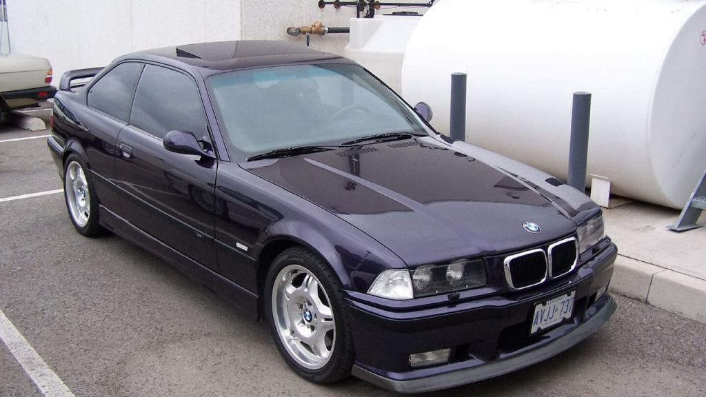 Dark purple BMW M3 E36 coupe with silver alloy wheels parked in concrete parking garage with white walls and pillars