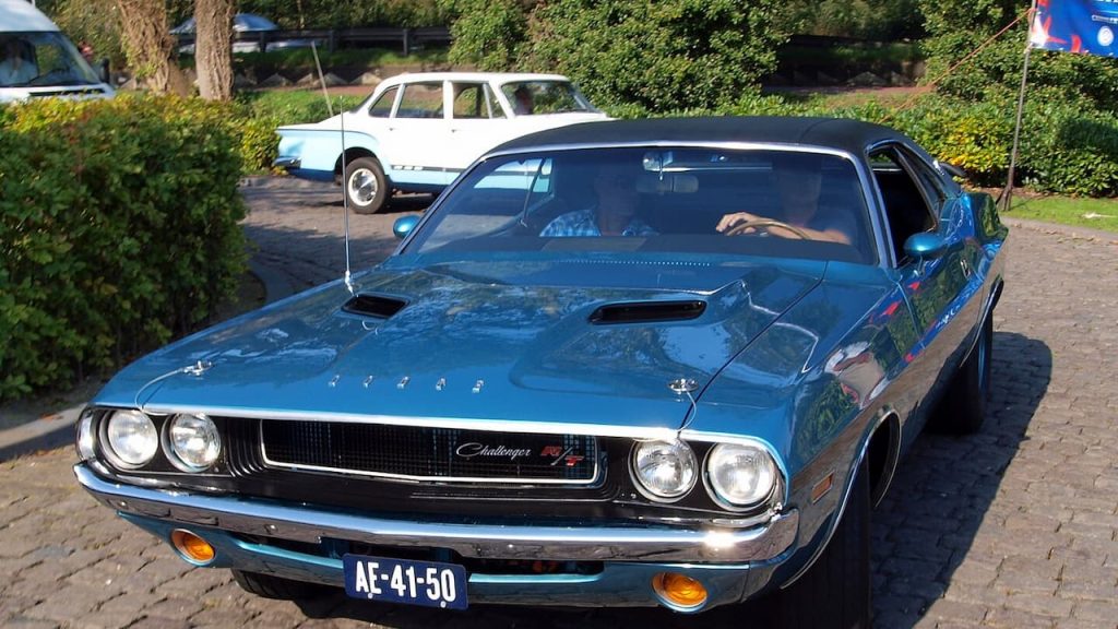 Blue 1970 Dodge Challenger muscle car parked on cobblestone with vintage white sedan visible in background
