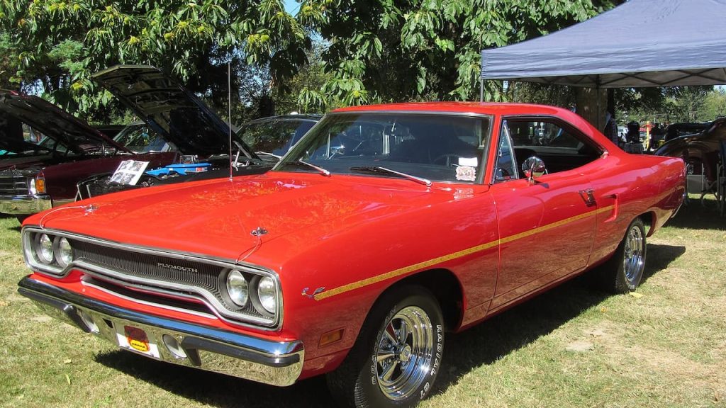 Bright red 1970 Plymouth Road Runner with yellow stripe displayed at outdoor car show under leafy trees