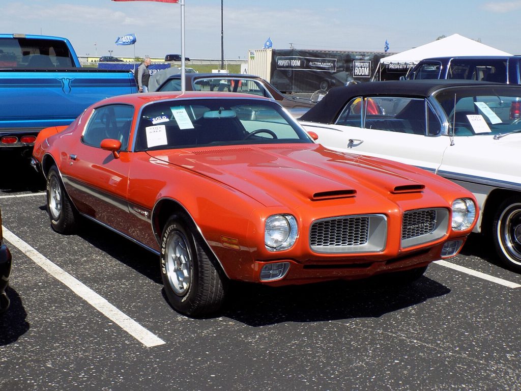 Bright orange 1972 Pontiac Firebird Formula 455 HO parked at a car show, surrounded by classic vehicles and spectators