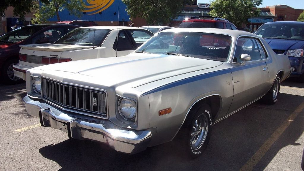 White 1976 Plymouth Volare Road Runner with blue racing stripes parked on street among other classic cars in urban setting
