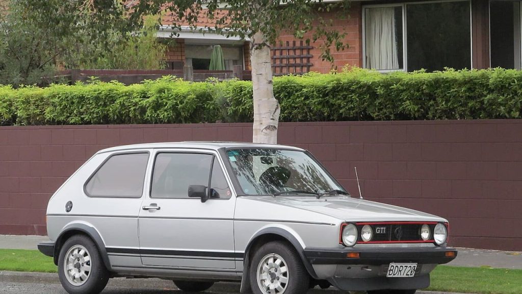 White vintage Volkswagen GTI Mk1 parked on residential street with brick wall, hedge, and birch tree behind