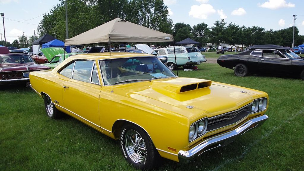 Bright yellow Plymouth GTX muscle car with hood scoop displayed at outdoor car show among other classic vehicles