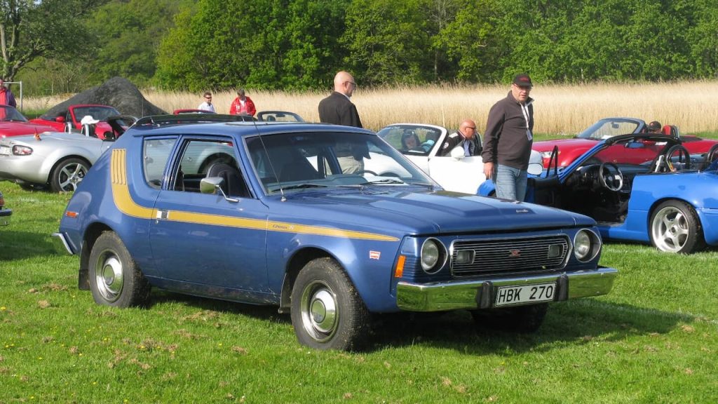 Blue AMC Gremlin with yellow racing stripes parked on grass at outdoor car show with other classic vehicles and spectators