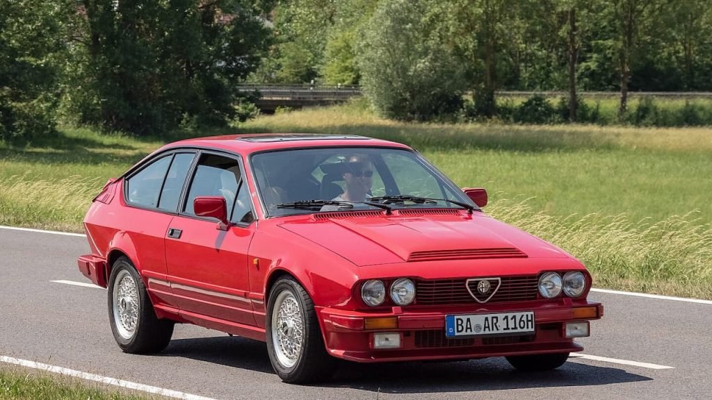 Red Alfa Romeo GTV6 with German license plate "BA AR 116H" driving on rural road surrounded by green countryside