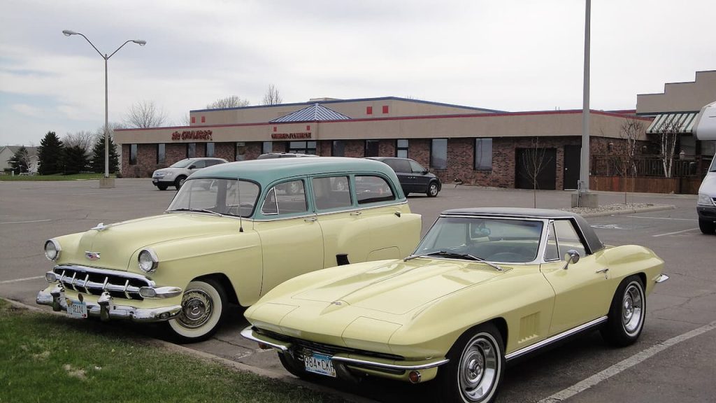 
Two pale yellow classic American cars - 1950s Chevrolet sedan and Corvette convertible - parked outside commercial building