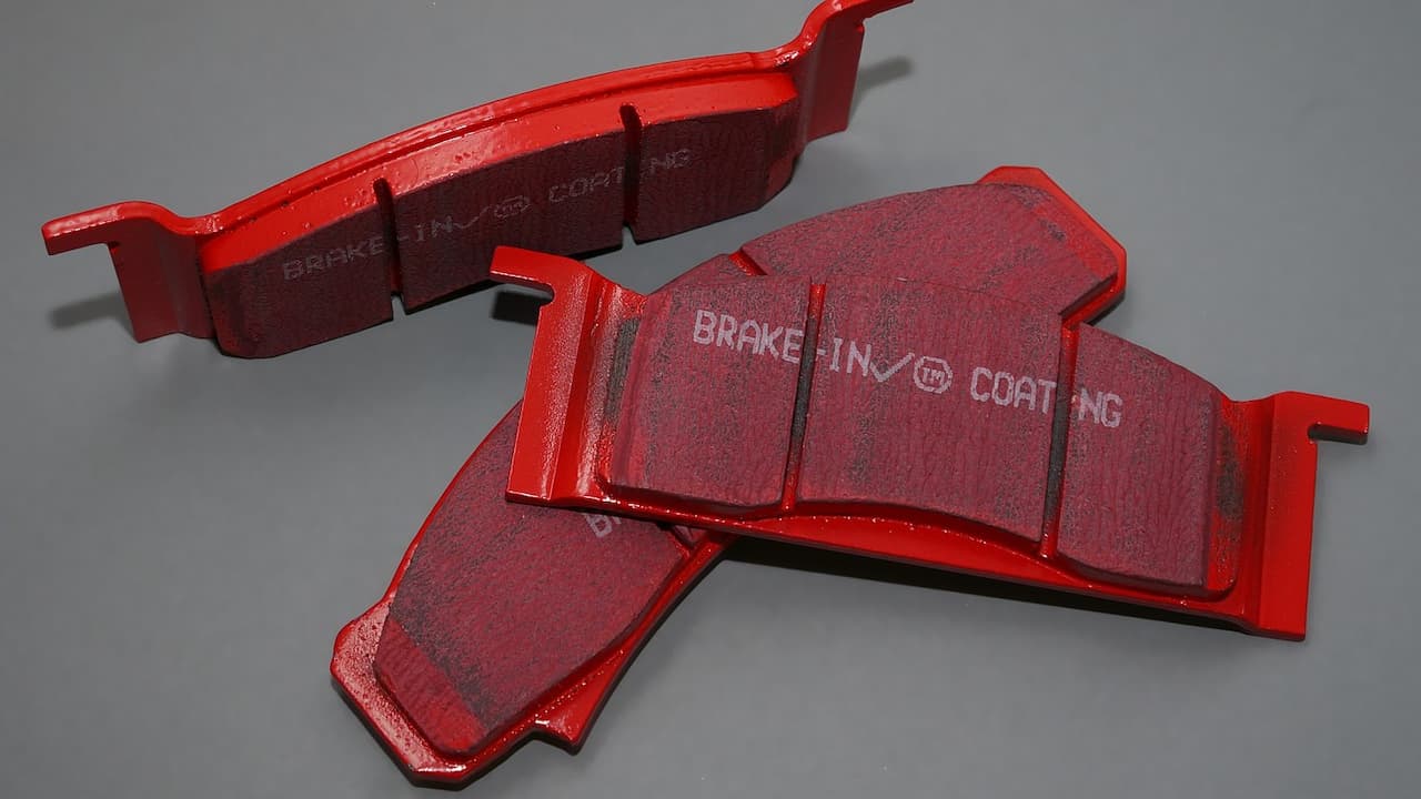 Red performance brake pads with "BRAKE-IN COATING" text, showing friction material surfaces and metal backing plates on gray background
