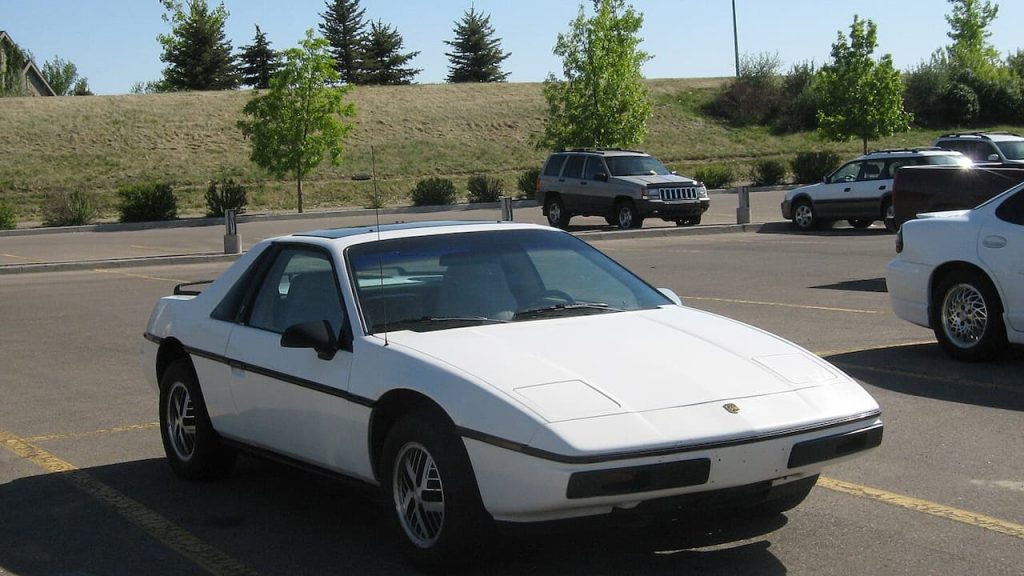 White Pontiac Fiero with black trim parked in lot surrounded by other vehicles and landscaped hillside