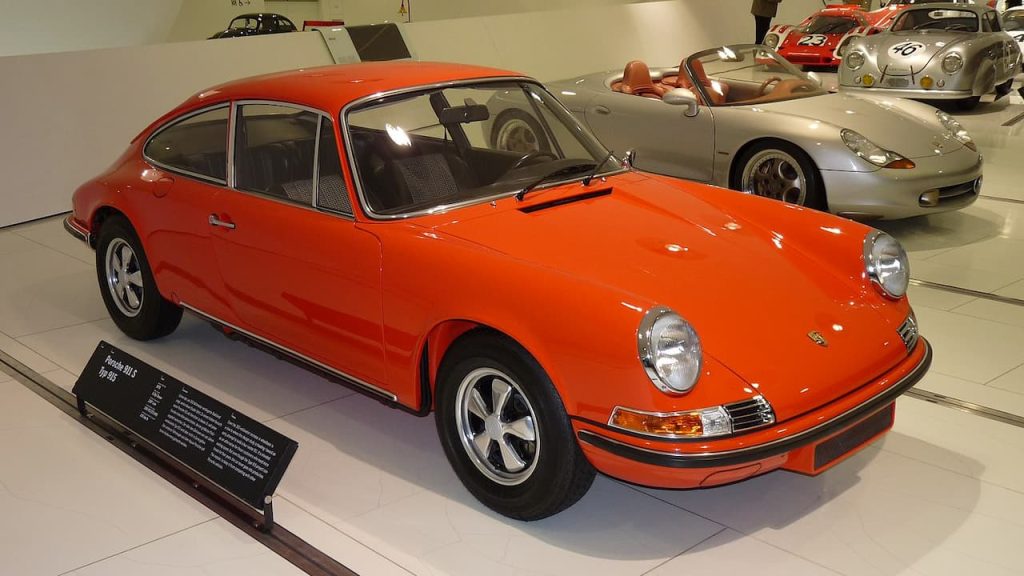 Orange vintage Porsche 911 sports car displayed in museum with silver convertible Porsche and other classic vehicles in background