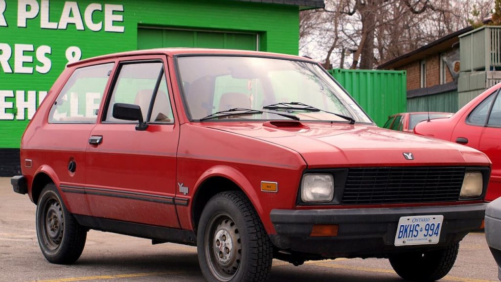 Red Yugo GV with Ontario license plate BKHS-994 parked in front of green commercial building and bare trees