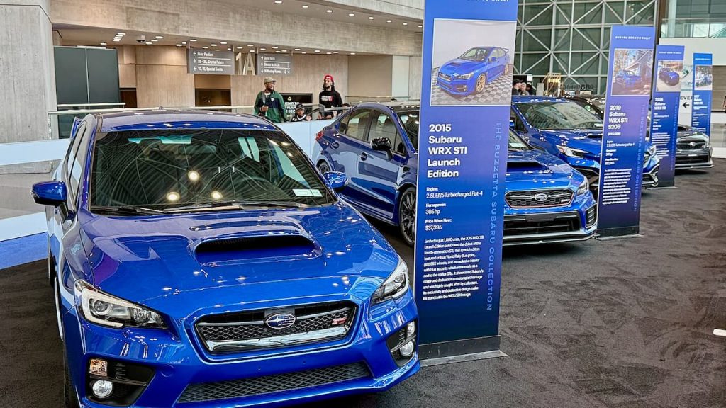 Blue Subaru WRX STI displayed at auto show with informational banner, surrounded by other vehicles in modern convention center