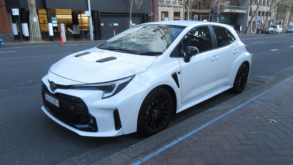 White Toyota GR Corolla with black wheels and sporty body kit parked on cobblestone street in urban setting