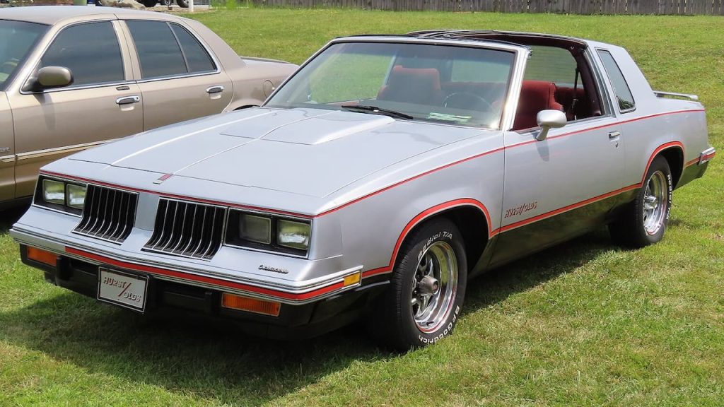 Silver 1984 Oldsmobile Hurst/Olds with red accent stripes and T-tops parked on grass at car show event