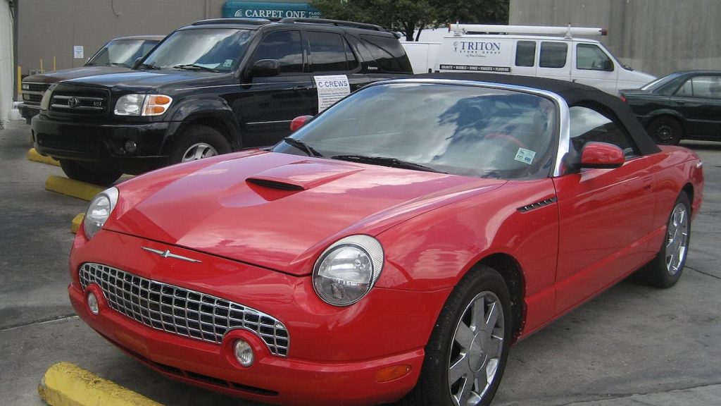 Red Ford Thunderbird convertible with retro styling and chrome wheels parked in commercial lot with SUVs and work vans