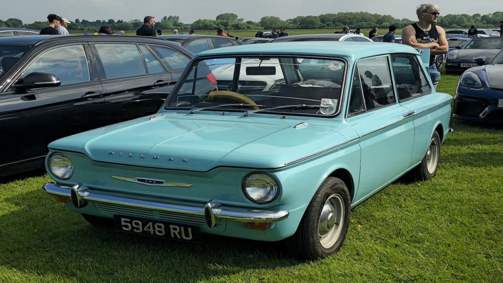 Light blue Hillman Imp classic car displayed at outdoor car show with other vintage vehicles and spectators on grass