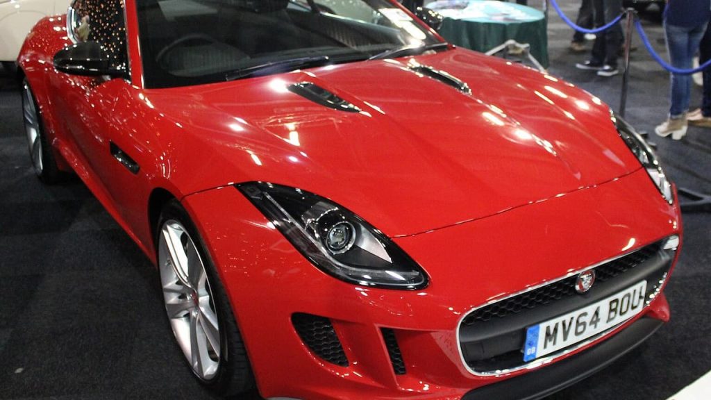 Red Jaguar F-Type sports car with UK license plate displayed at indoor automotive exhibition with visitors nearby