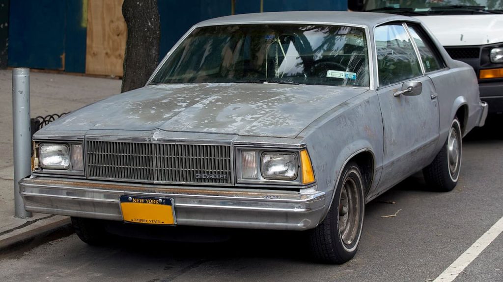 Weathered gray 1980 Chevrolet Malibu with New York license plate parked on city street showing signs of age