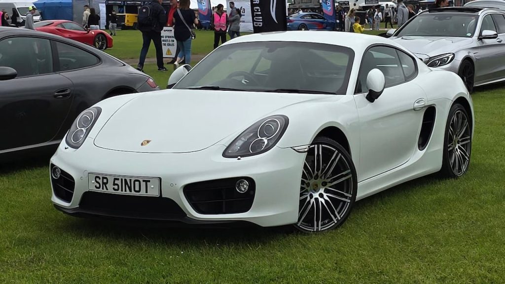 White Porsche Cayman sports car with license plate "SR 51NOT" displayed on grass at outdoor car show event