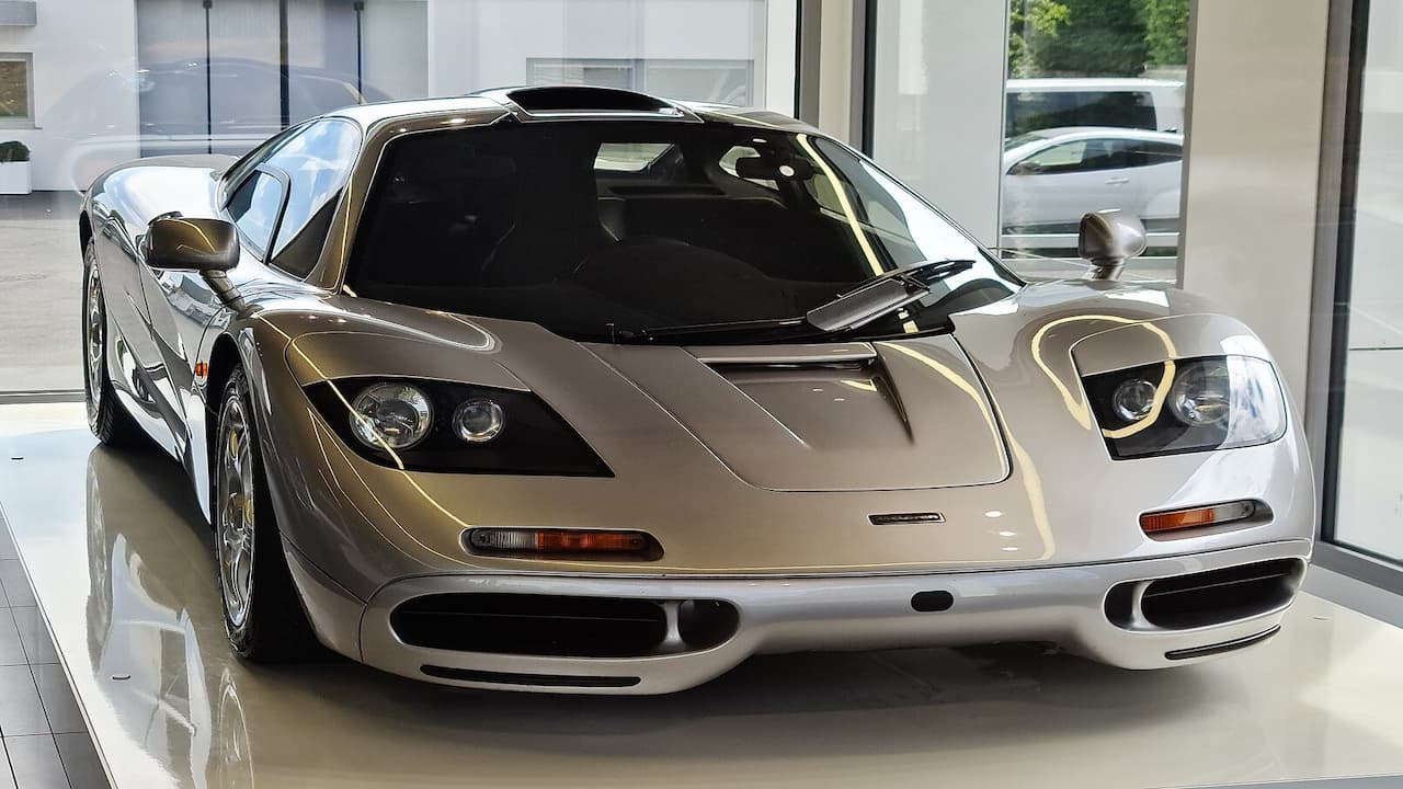 Alt description (20 words): Silver McLaren F1 supercar with distinctive center-positioned driver's seat displayed in glossy showroom with glass walls