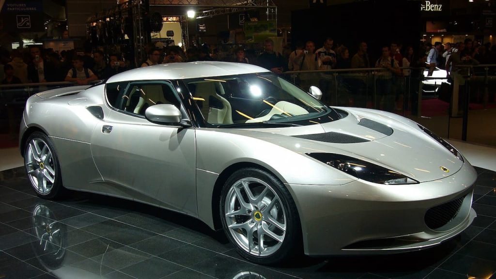 Silver Lotus Evora sports car displayed at auto show with crowds of spectators behind barriers watching the vehicle presentation