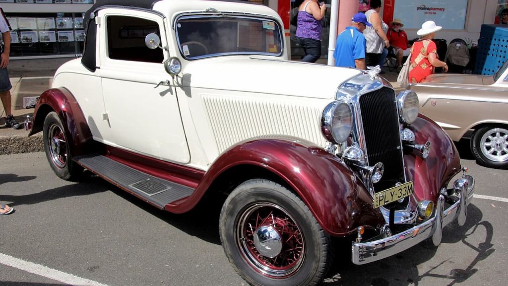 Vintage cream and burgundy 1933 Plymouth Coupe with PLY-33M plates at car show, featuring chrome trim and wire wheels
