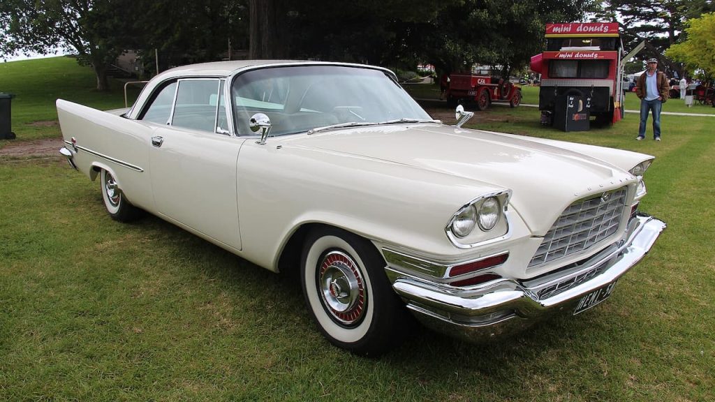 A white 1958 Chrysler 300D hardtop coupe with chrome trim, parked on grass near a red food stall and trees