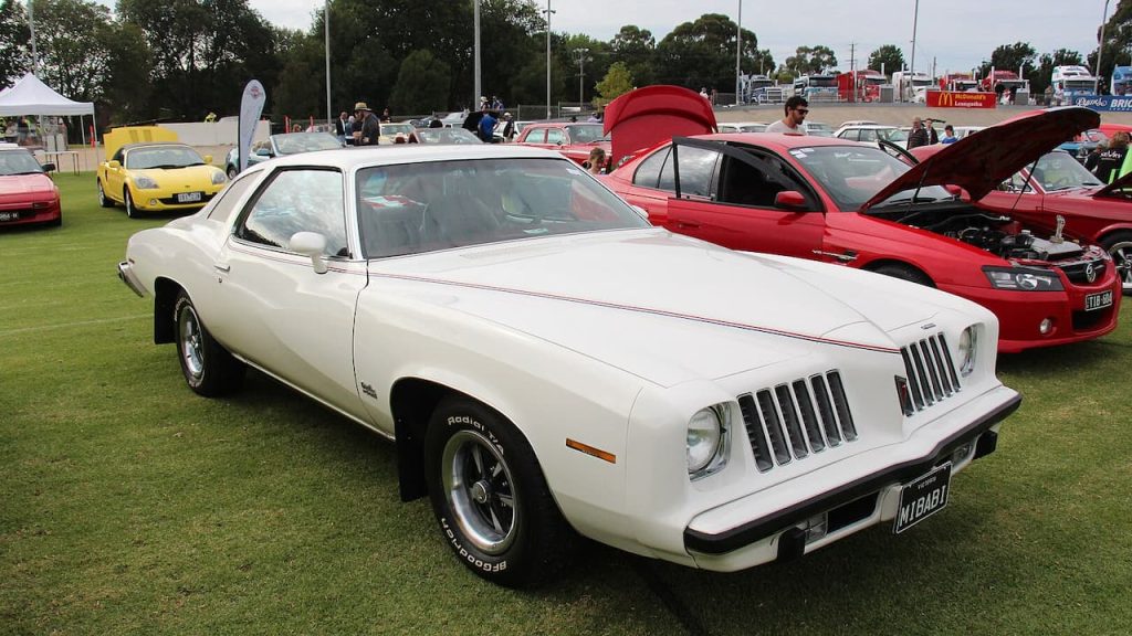 White 1974 Pontiac 4-4-2 Cutlass muscle car displayed at outdoor car show with other classic vehicles on grass field