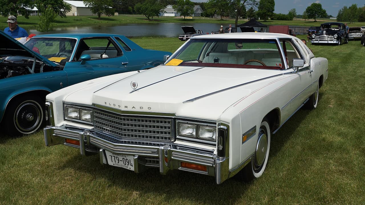 White 1978 Cadillac Eldorado with Minnesota license plate displayed at lakeside car show alongside blue classic car