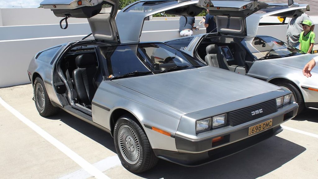 Silver DeLorean DMC-12 with signature gull-wing doors open, displaying black interior on concrete surface with spectators observing nearby