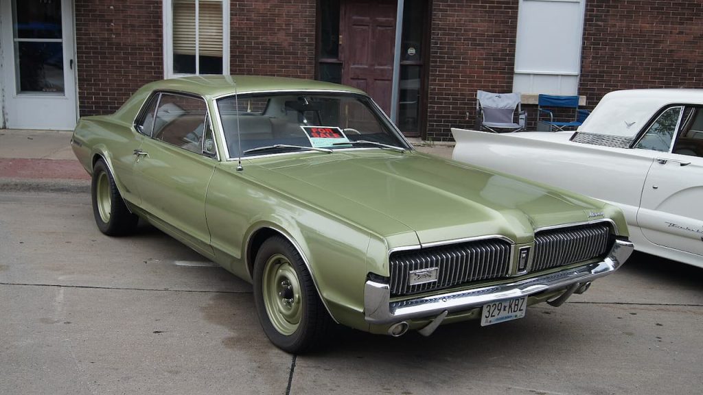 Green Mercury Cougar classic car with chrome grille and steel wheels parked on concrete near brick building