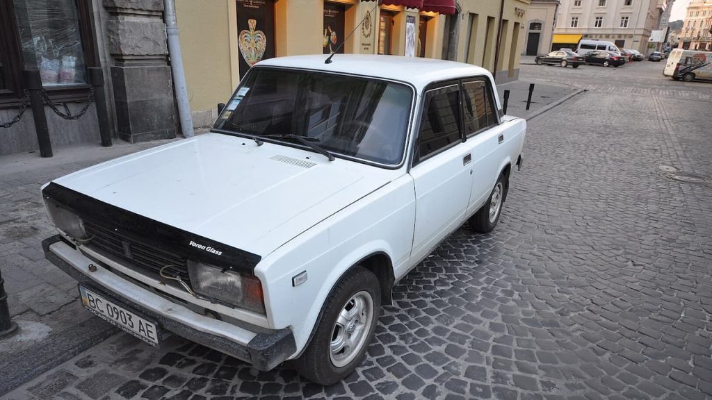 White Lada Riva parked on cobblestone street in European city center with historic buildings and storefronts behind