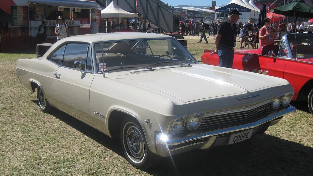 White vintage 1965 Chevrolet Impala Super Sport Coupe with "COUPE" license plate displayed at outdoor classic car show with spectators and tents