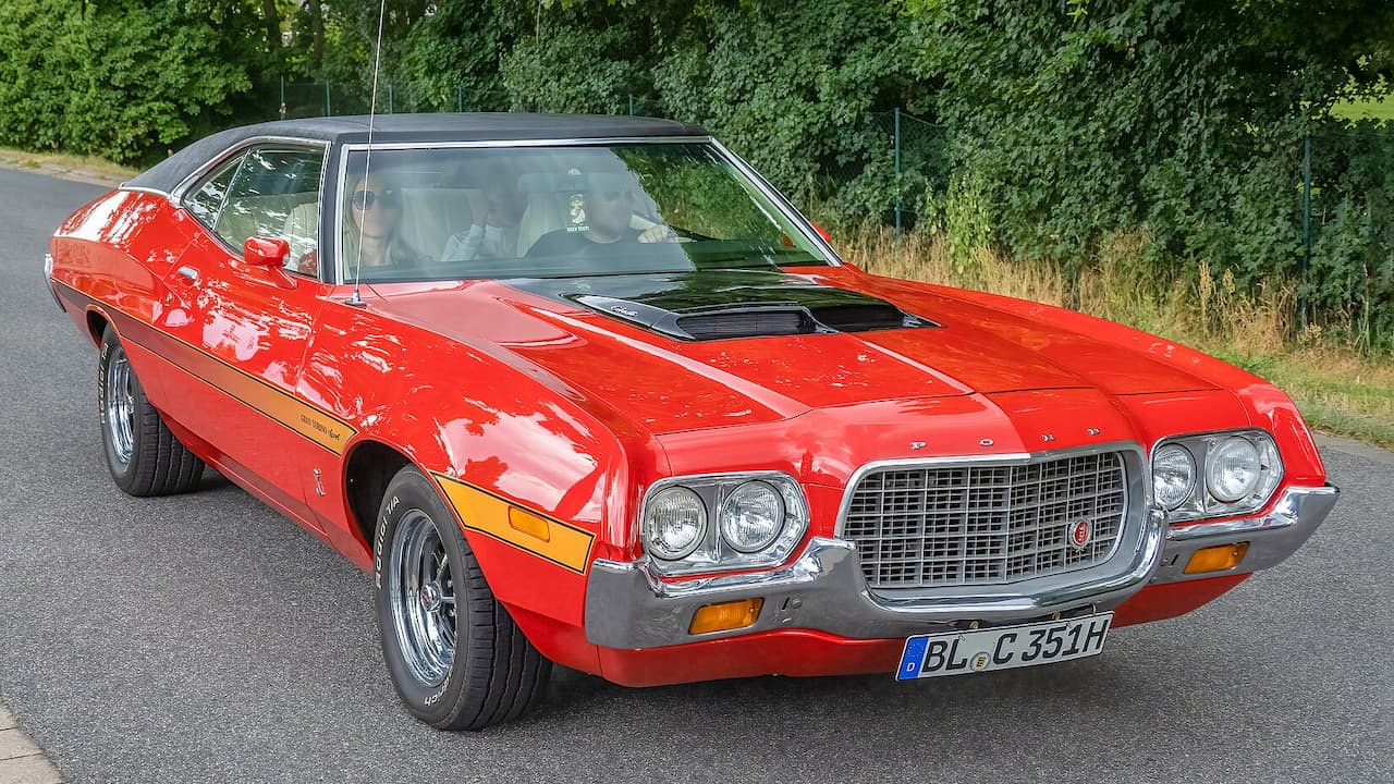 Red Ford Gran Torino with black roof and hood scoop driving on road with German license plate and occupants inside