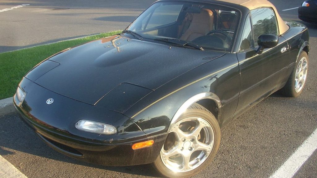Black Mazda Miata with tan soft top and silver alloy wheels parked on street beside grass area