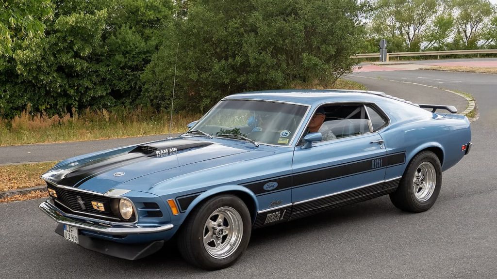 Classic blue Ford Mustang Mach 1 with black racing stripes parked on rural road surrounded by green trees