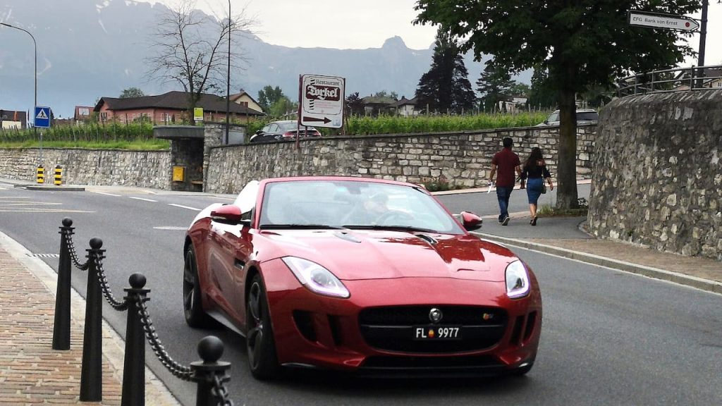 Red Jaguar F-Type R parked on street with stone wall and mountain backdrop visible