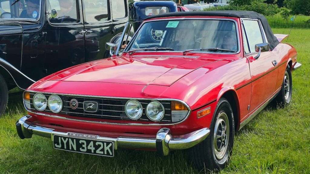 Red Triumph Stag convertible with license plate JYN 342K parked on grass at classic car show with other vintage vehicles