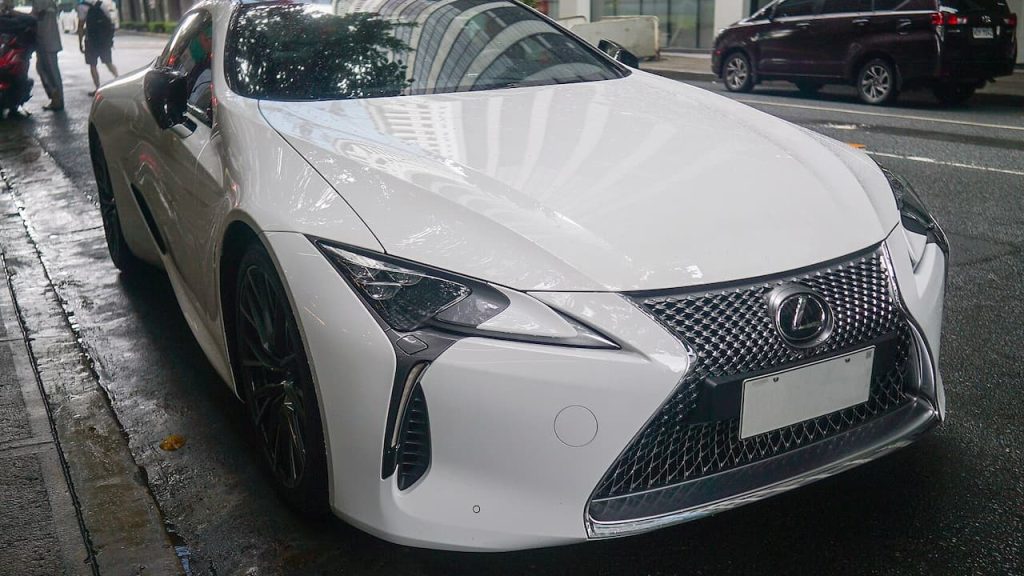 White Lexus LC 500 with distinctive spindle grille and angular headlights parked on wet urban street