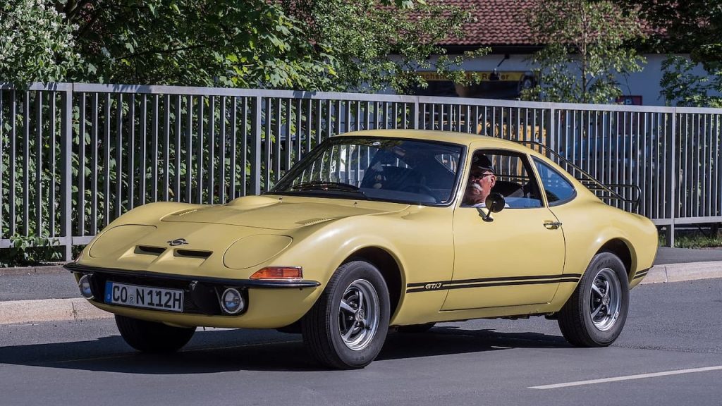Yellow Opel GT classic sports car with driver, parked on street beside metal fence with trees and house behind