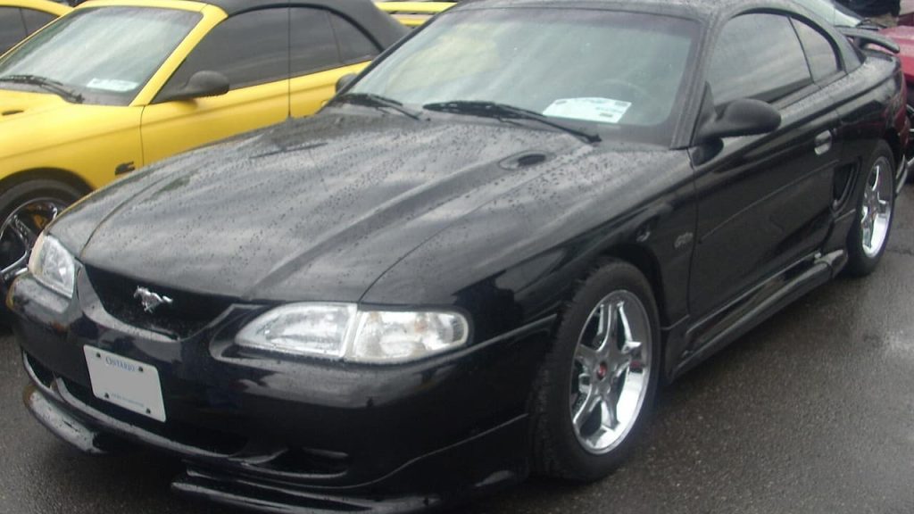 Black tuned Ford GT sports car parked on asphalt with yellow vehicle visible in background at car meet