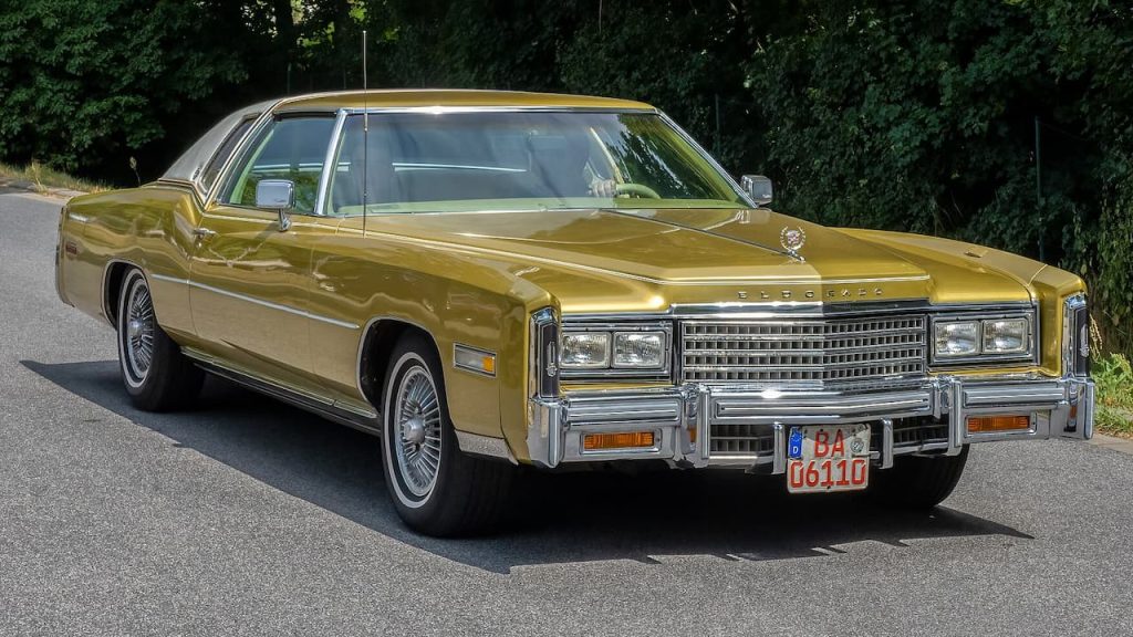 Gold Cadillac Eldorado classic luxury coupe with chrome bumpers and wire wheels parked on asphalt road near tree