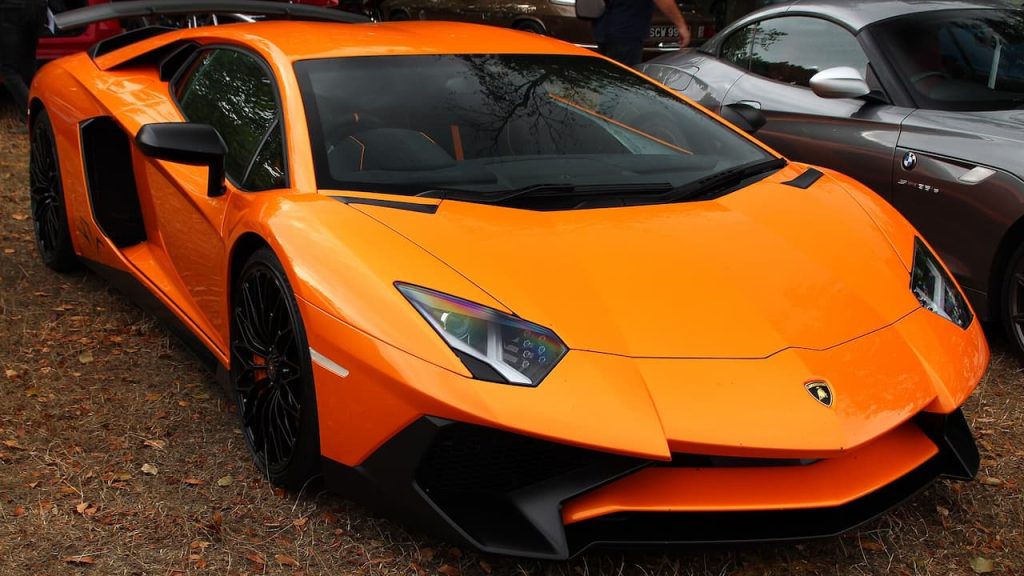 Bright orange Lamborghini Aventador SV supercar with black wheels and angular design parked among other vehicles outdoors