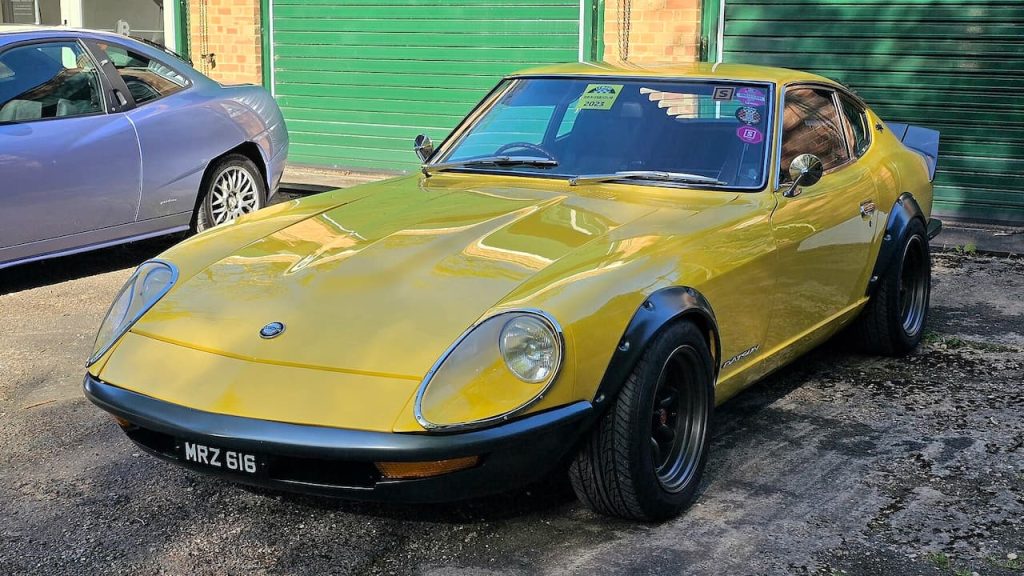 Yellow Datsun 240Z with license plate "MRZ 616" parked on street beside green roller shutters and gray sedan