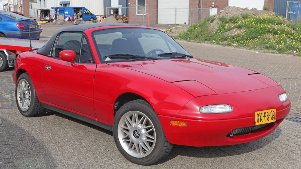  Red Mazda MX-5 Miata convertible with black soft top parked on cobblestone street in urban industrial area setting