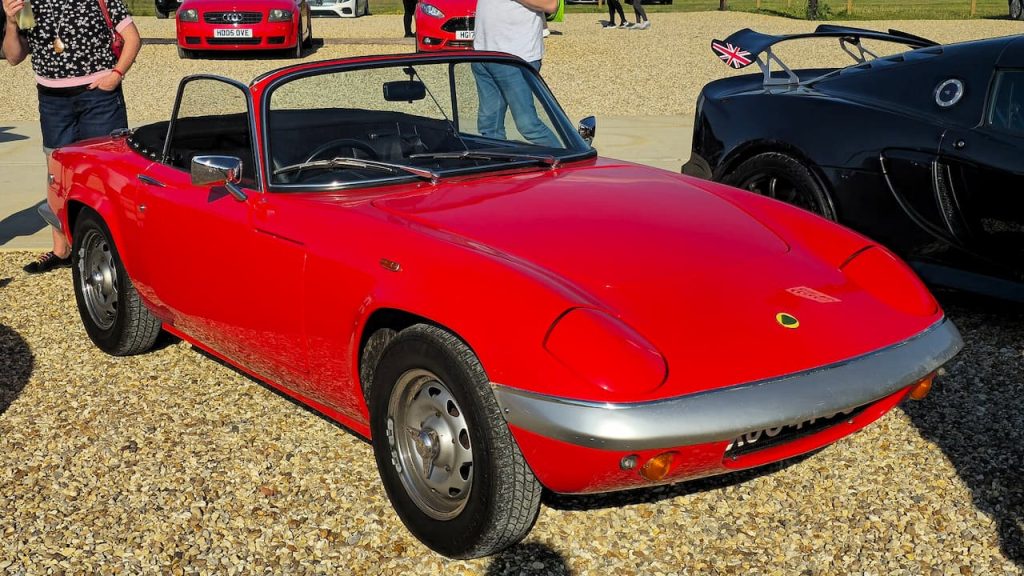 Red Lotus Elan convertible sports car with black soft top and steel wheels at outdoor car show on gravel
