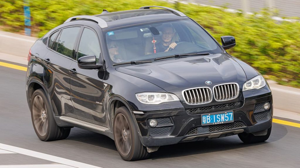 Black BMW X6 with license plate B-ISW57 driving on road with motion blur and green foliage background